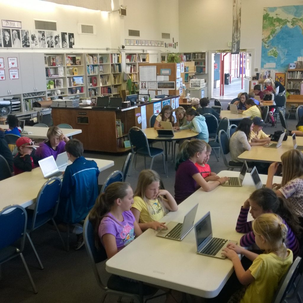Students At Tables In Library. They Are Working With Their Partner On Hour Of Code.